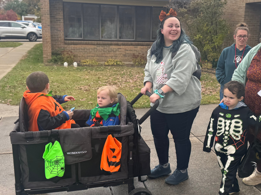 Adult with blue hair pushing a wagon with two small children dressed as pumpkins. There is also a small child in a skeleton costume, with two, partially in-frame adults.
