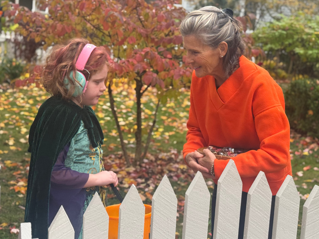 Young student with short, curly hair, dressed as Merida from Brave receives candy from a woman wearing a bright orange sweater while trick-or-treating.