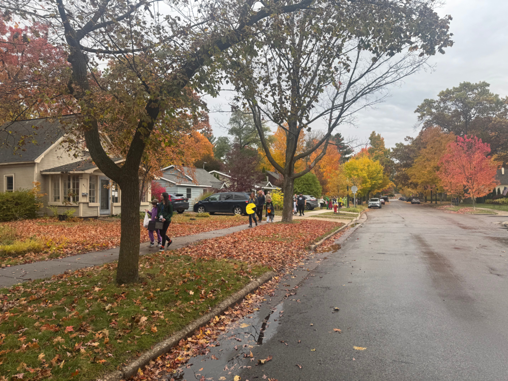 Image of the neighborhood while trick-or-treating. It is overcast, the ground is wet, with bright green, red and orange leaves in the trees and on the ground.