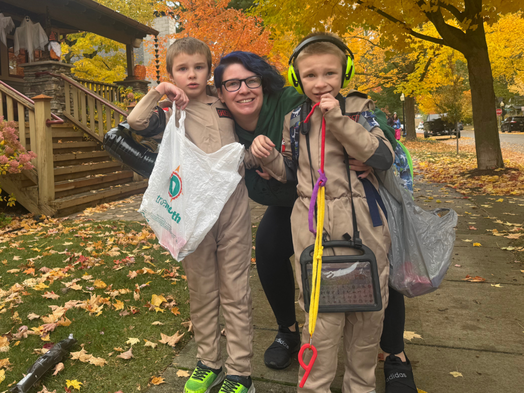 Image of parent with blue hair posing and smiling between their two boys, dressed up as Ghost Busters while trick-or-treating.