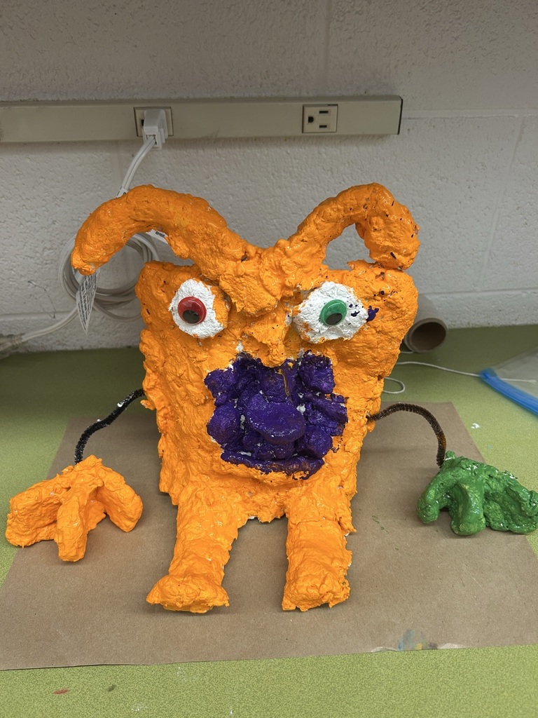 Image of an orange creature with a purple mouth, floppy antenna, one orange hand, one green hand, one red eye, and one green eye, made with paper mache.