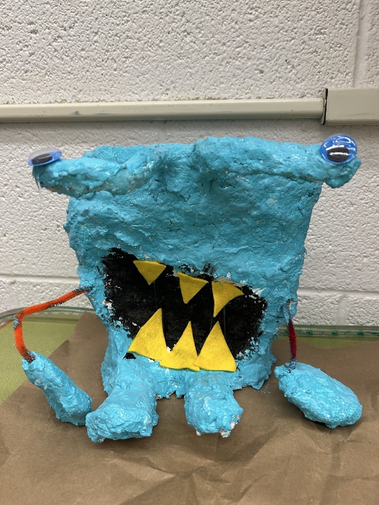 Image of a blue paper mache creature, with small eyes on either side of its rectangular head and sharp, yellow teeth.