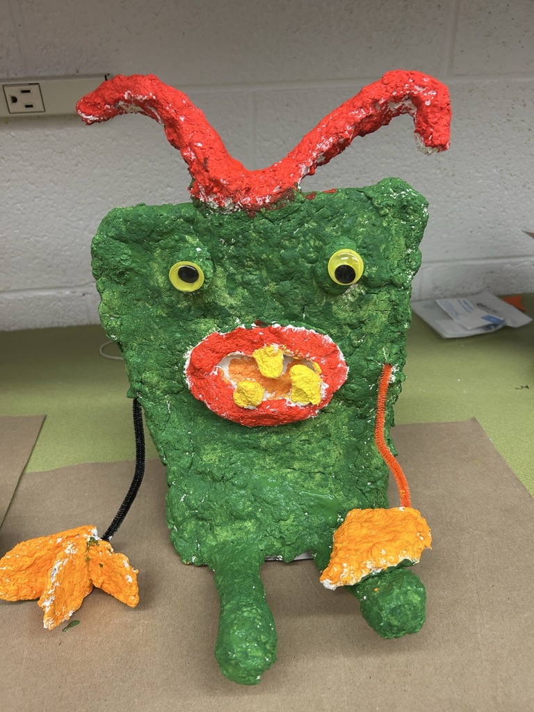 Image of a green paper mache creature, with floppy red antennae, small yellow eyes, red mouth, orange tongue, three yellow teeth, and orange hands.