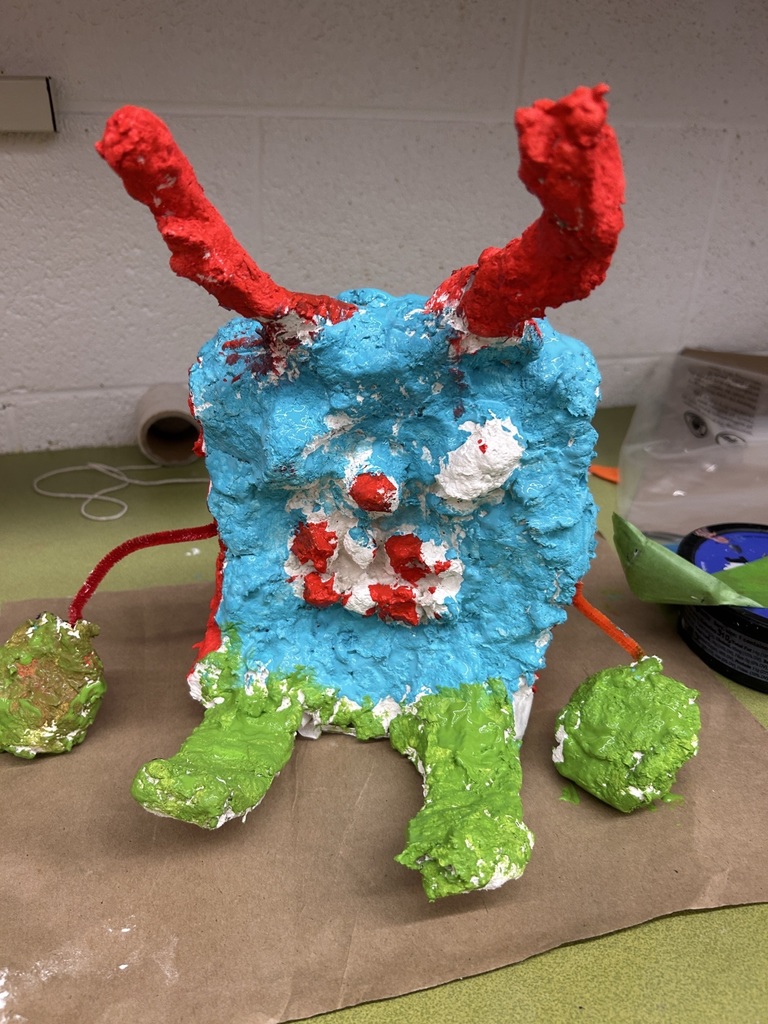 Image of a blue creature with red antenna, green limbs, and several red, indistinguishable facial features, made with paper mache.