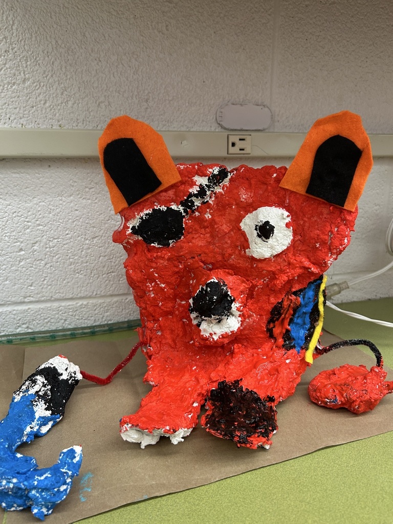 Image of a red paper mache creature, with orange and black felt ears. An eye patch, one red hand, a blue hook in place of a hand, black nose, one red foot and one black foot.