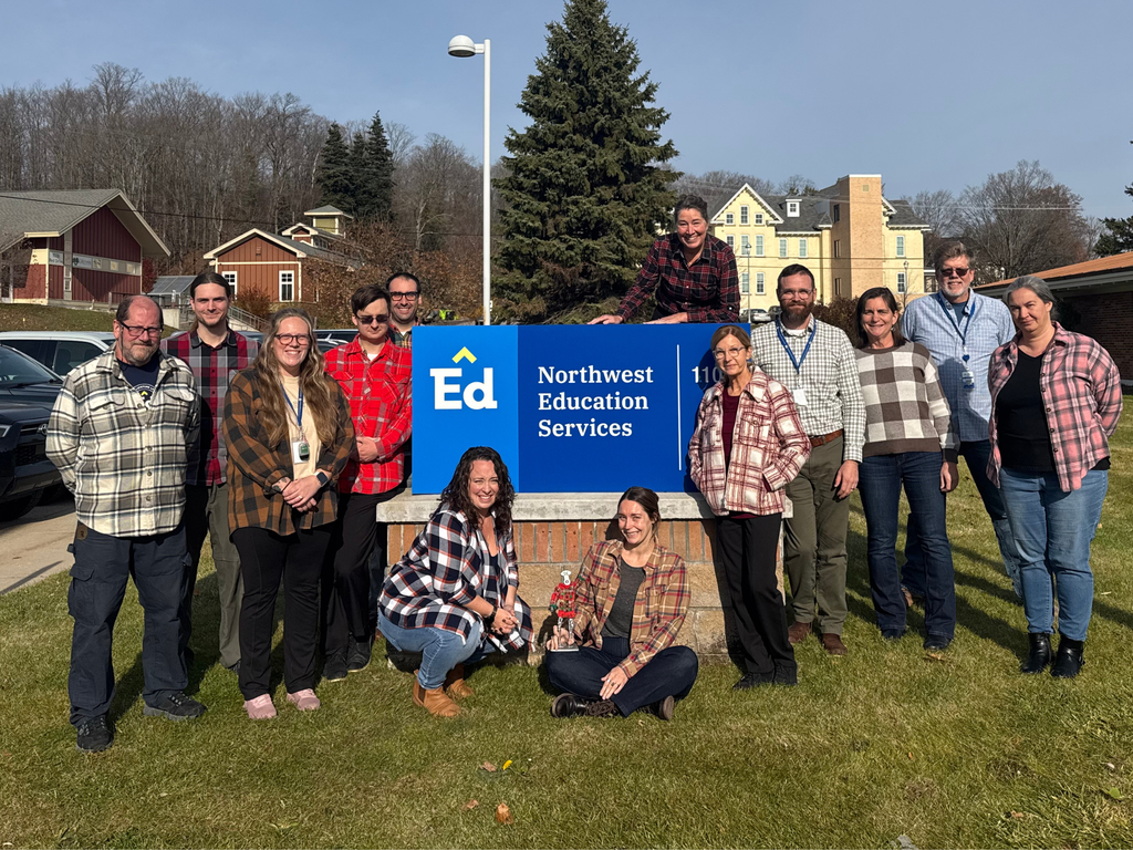 A group of staff members wearing flannel shirts stand outdoors around a blue Northwest Education Services sign. They are smiling and posing on a grassy area with buildings and trees visible in the background under a clear sky