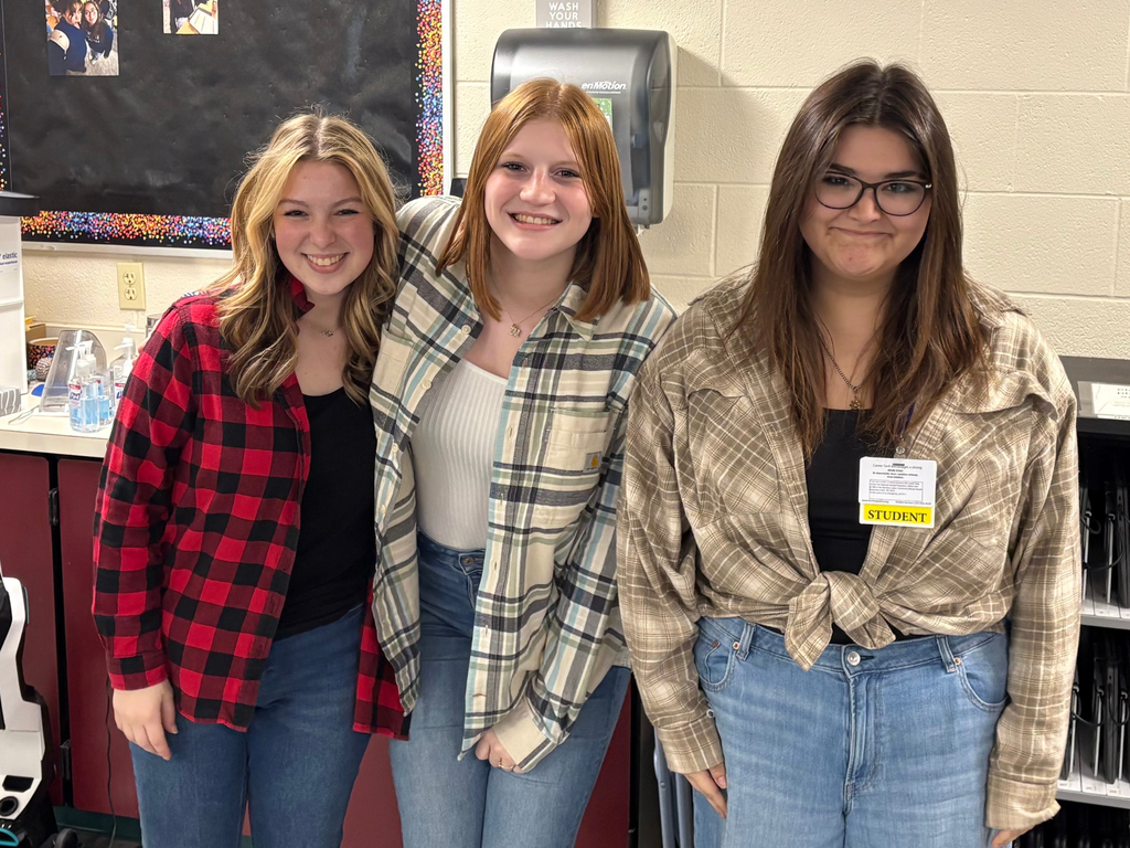 Three students stand together in a classroom space, all wearing different styles of flannel shirts. They are smiling in front of a bulletin board and classroom supplies.