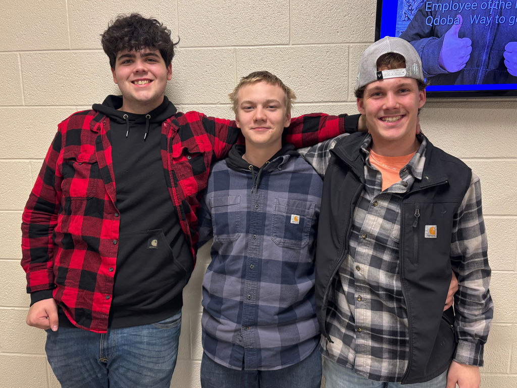 Three students stand close together in front of a light-colored brick wall. They are all wearing flannel shirts and smiling, with one student’s arms around the others.