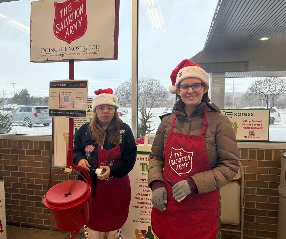 Two volunteers wearing red Salvation Army aprons and Santa hats stand inside a grocery store entrance next to a red Salvation Army donation kettle on a stand. Both volunteers hold a bell and smile toward the camera. Salvation Army signage is displayed above them, with additional store signs, large windows, and a snowy parking lot visible in the background. 