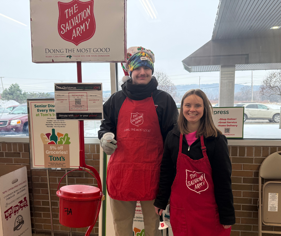Two smiling volunteers wearing red Salvation Army aprons stand inside the entrance of a grocery store next to a red Salvation Army donation kettle on a stand. Both volunteers stand side-by-side holding a small bell. Behind them are Salvation Army signs and store notices, with large windows showing a parking lot and winter scenery outside.