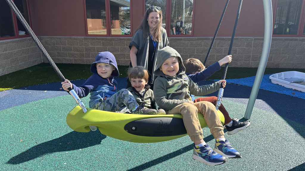 A group of 5 children smiling on a large swing set, being pushed by a parent. 
