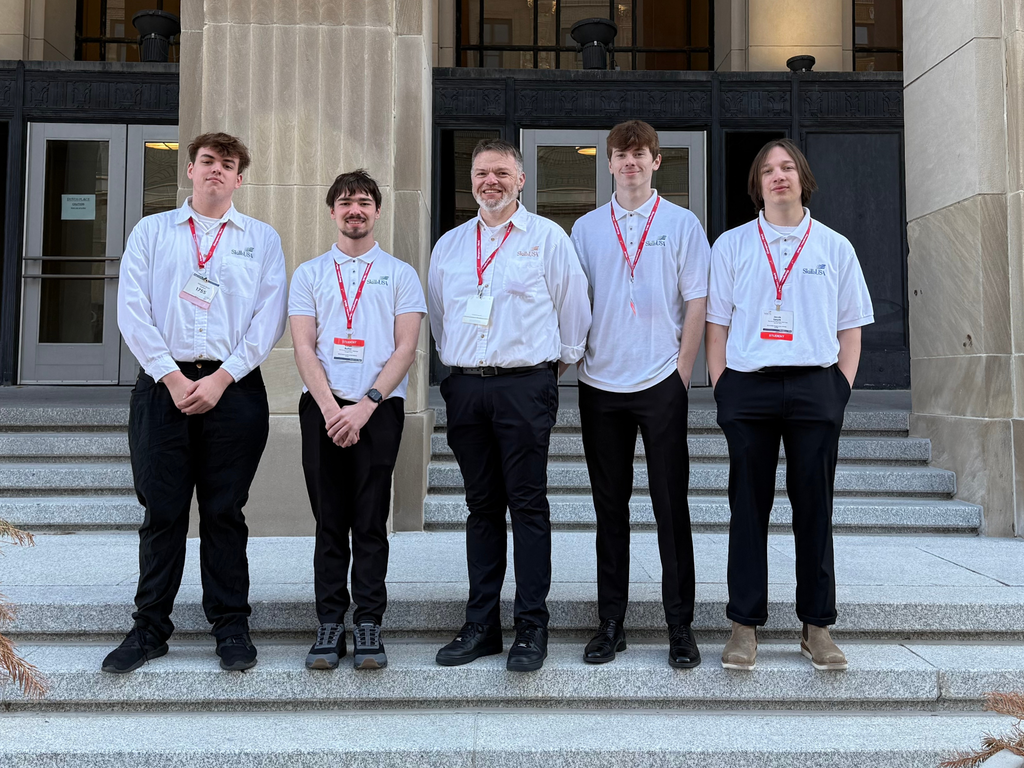 Image of Jake Smyth, Cohl Jennings, George Shumar, Tyler Evernham and Ethan Butler standing on exterior steps of the 2026 SkillsUSA Michigan venue, wearing white shirts, black pants and red lanyards, posing and smiling for a group photo. 