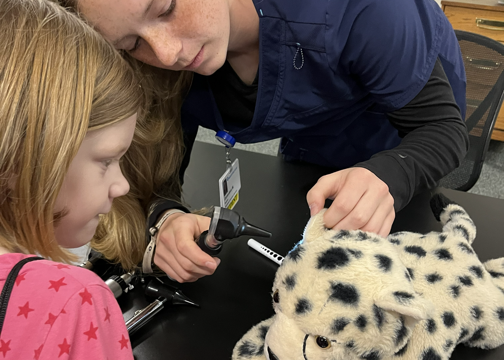 Picture of Career Tech Health Sciencies student wearing scrubs, using an otoscope to look into the ear of a elementary student’s stuffed animal. 