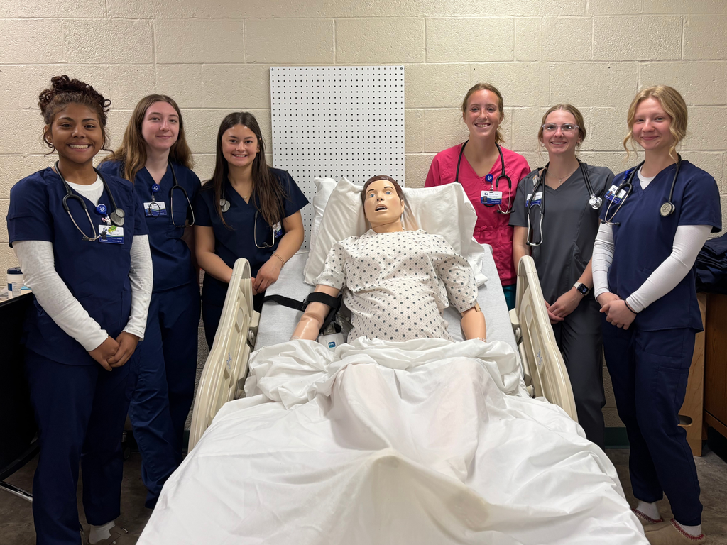 Six Career Tech Health Sciences students posing with a practice manikin placed in a hospital bed. 