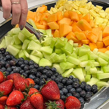 A hand holding tongs grabbing from a platter of pineapples, melons, blueberries and strawberries. 