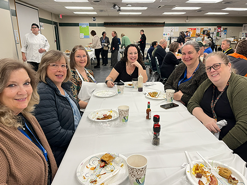 A group of North Ed staff members sitting around a long table, smiling for a group photo while eating breakfast. 
