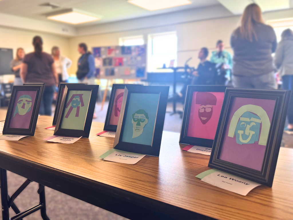 Photo: Framed self portraits made from brightly colored construction paper on a table with gallery attendees in the background. 