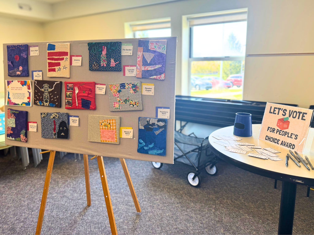 Photo: Fiber art created by multiple ACE students displayed on a pin board, next to a small round table with pens, paper ballots, ballot box and a sign that reads “Let’s vote for people’s choice award.”