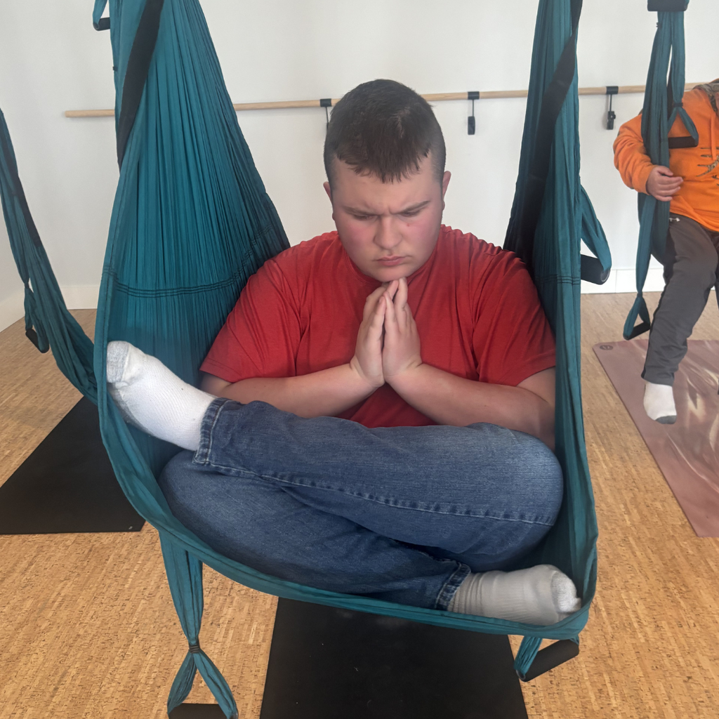 A student wearing a red shirt and jeans sitting in a trapeze swing with their legs crossed, eyes closed and head bowed in deep concentration. 
