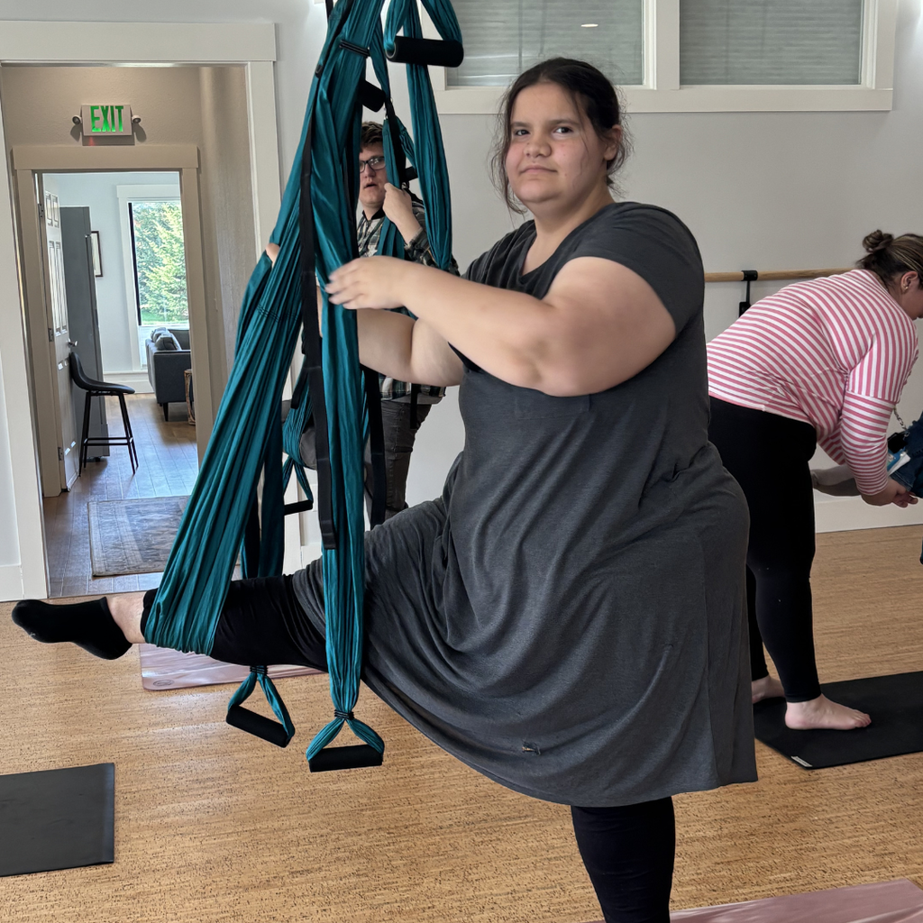 A student wearing a grey dress and black leggings holding on to a trapeze apparatus, standing  with one leg suspended in by the apparatus. 