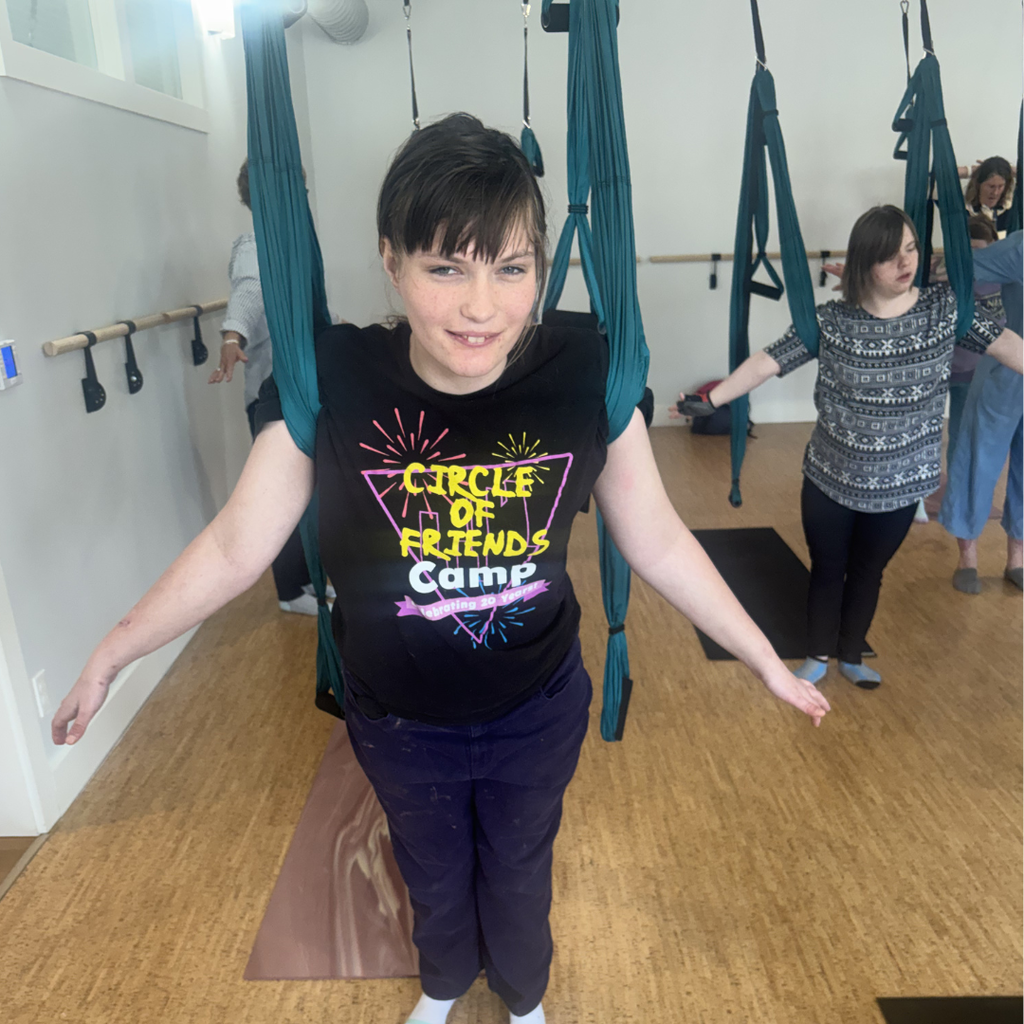 A student wearing a black graphic t-shirt and dark pants standing while being supported by a trapeze apparatus around their shoulders. 
