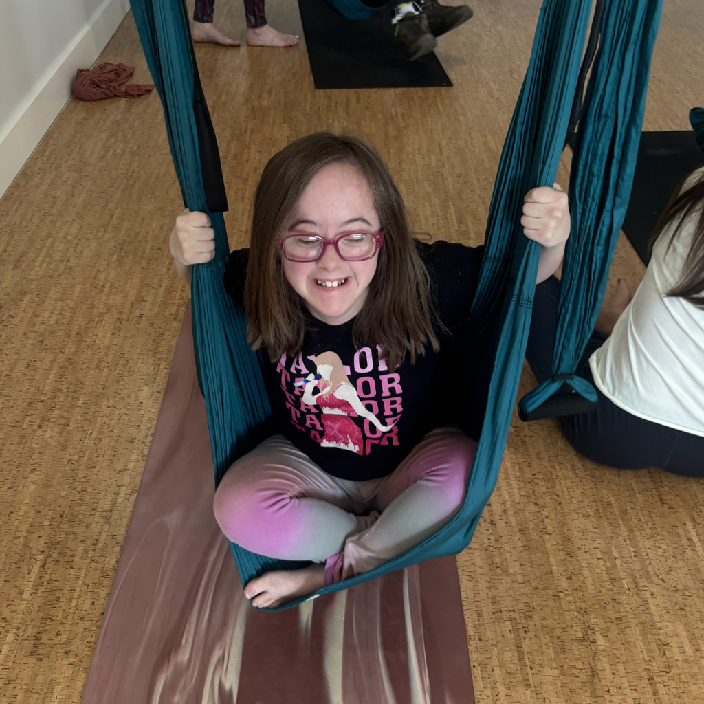 A student wearing a graphic t-shirt, tie-dye pants and sparkly glasses sitting with their legs crossed in a trapeze swing. 