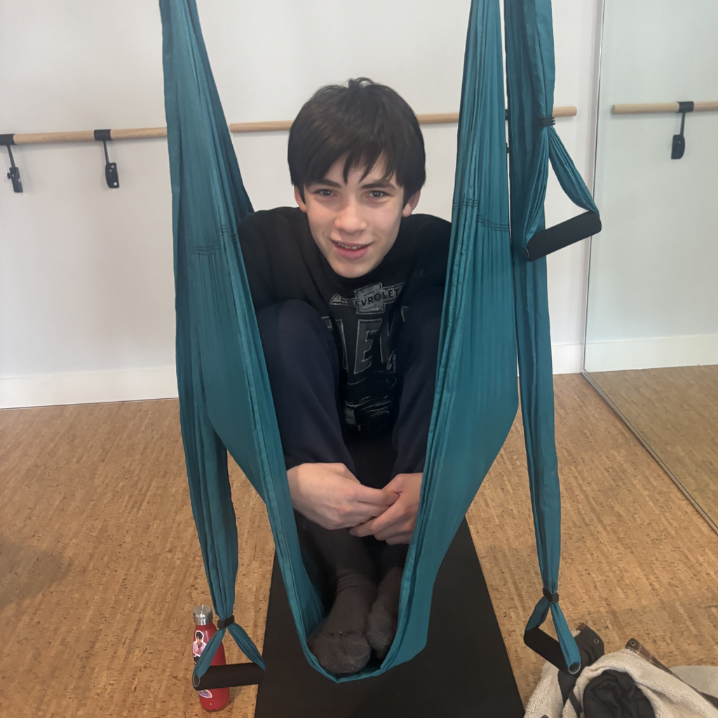A student wearing a dark shirt and dark pants, sitting in a trapeze swing, holding their legs close to their body, smiling at the camera. 