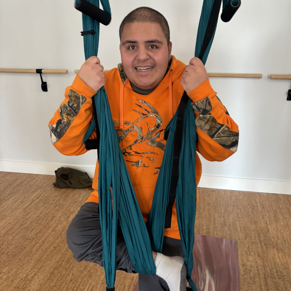 A student wearing an orange hoodie sitting in a trapeze swing smiling at the camera. 