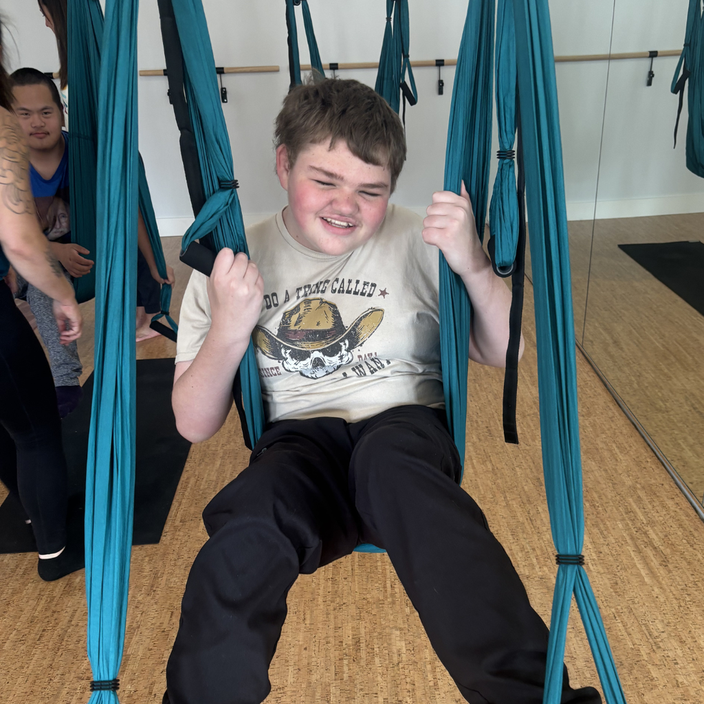 A student wearing a graphic t-shirt and black pants sitting in a trapeze swing, smiling. 