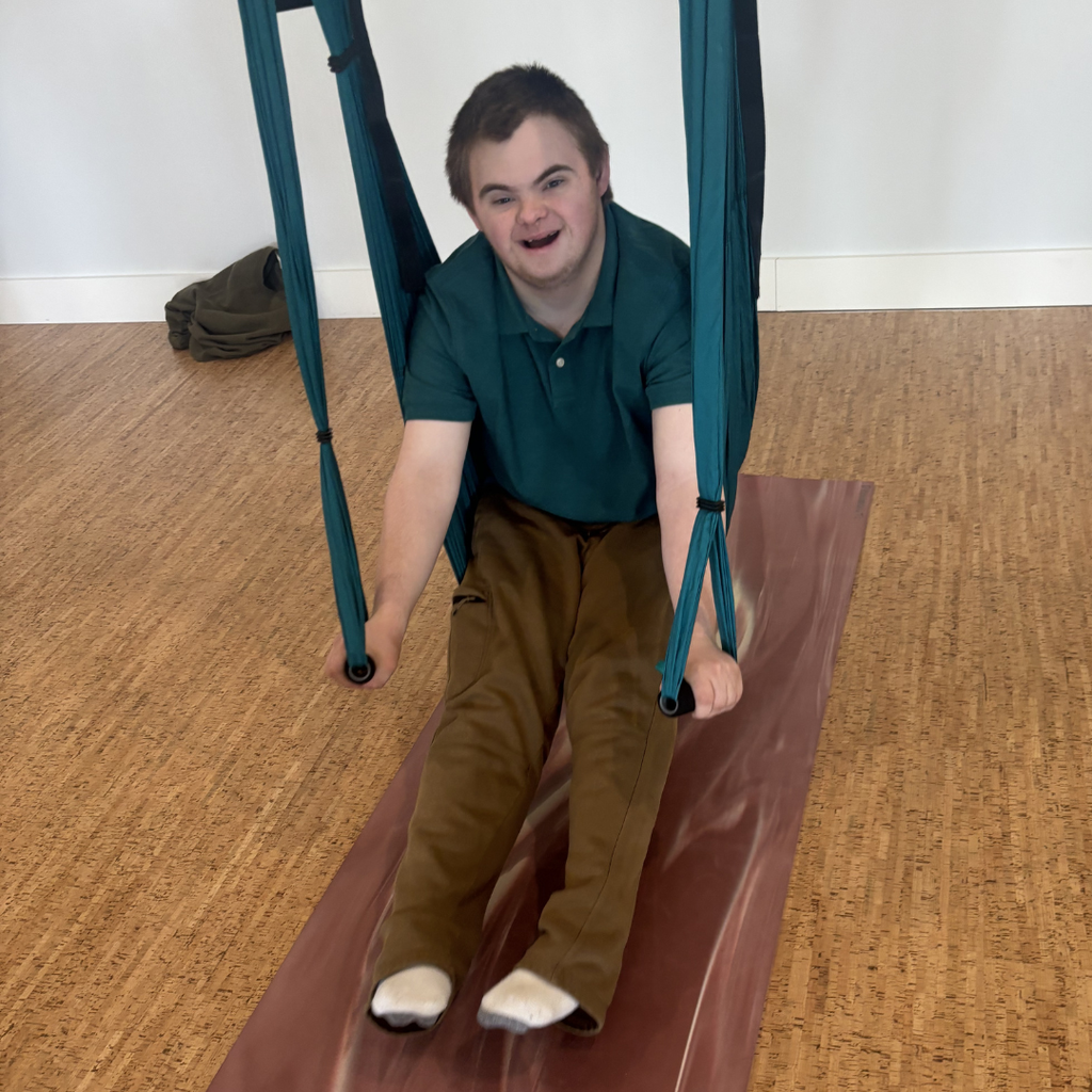 A student wearing a dark green polo and khaki pants sitting in a trapeze swing with their legs straight and feet on a yoga mat. 