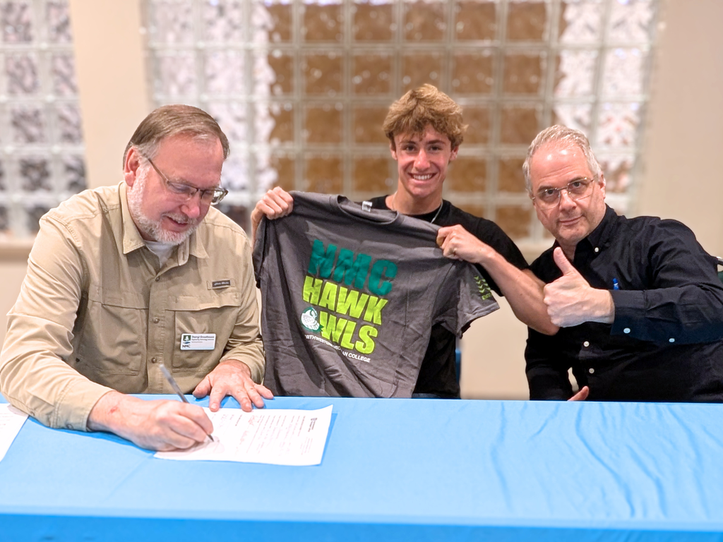 North Ed Career Tech student holding up an “NMC Hawk Owls” t-shirt, posing with their instructor who is giving a thumbs-up while a representative from Northwestern Michigan College signs the student’s articulation agreement.