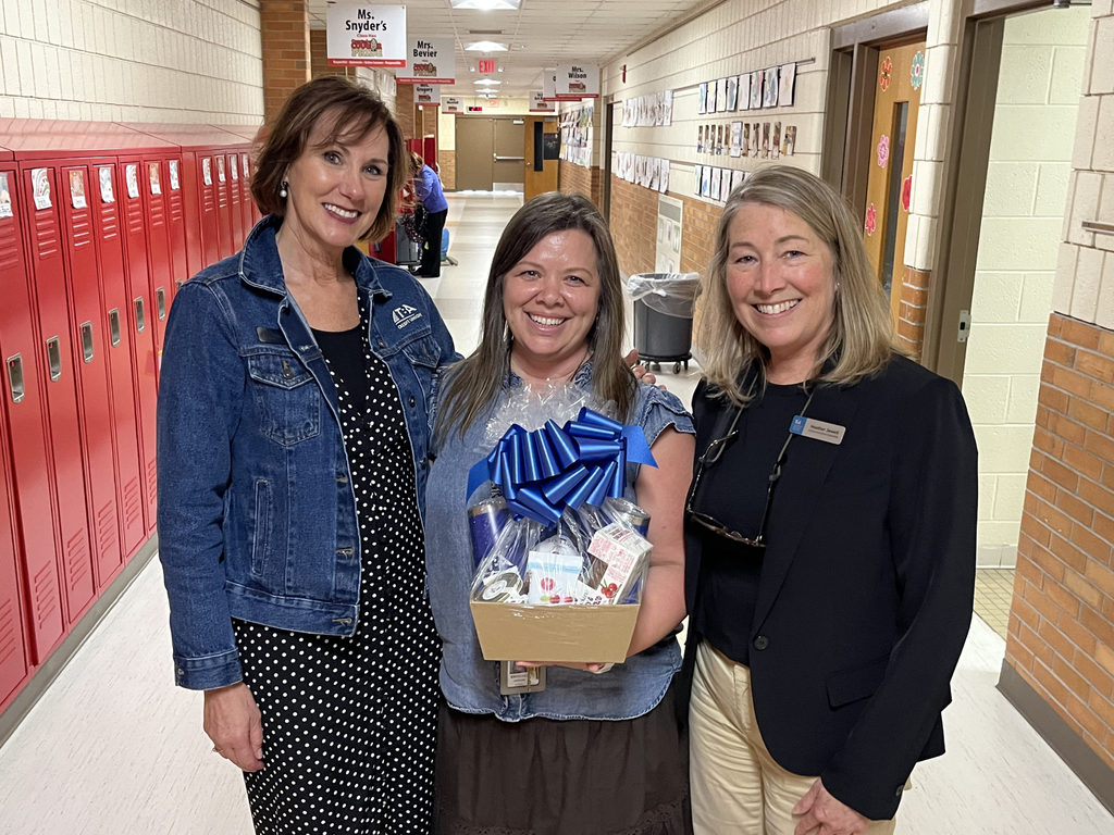 Kim Herron posing between Mary Beth Ruskowski and Heather Jewell, standing in the hallway at Courtade Elementary School, holding a gift basket with a large blue ribbon. 
