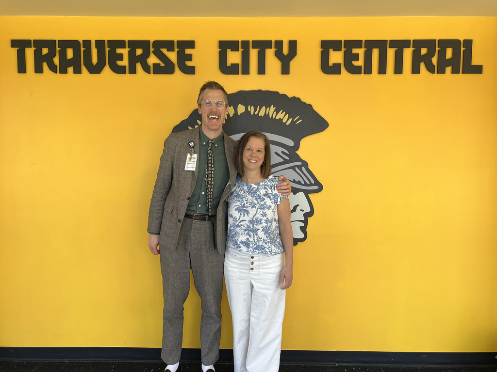 Teacher Becky Harvey and School Principal Ben Berger smiling and standing in front of a yellow wall painted with the Traverse City Central logo. 