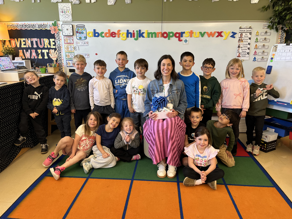 Jessica Lawrence posing with her students while sitting in a chair holding a gift basket with a large blue ribbon. 