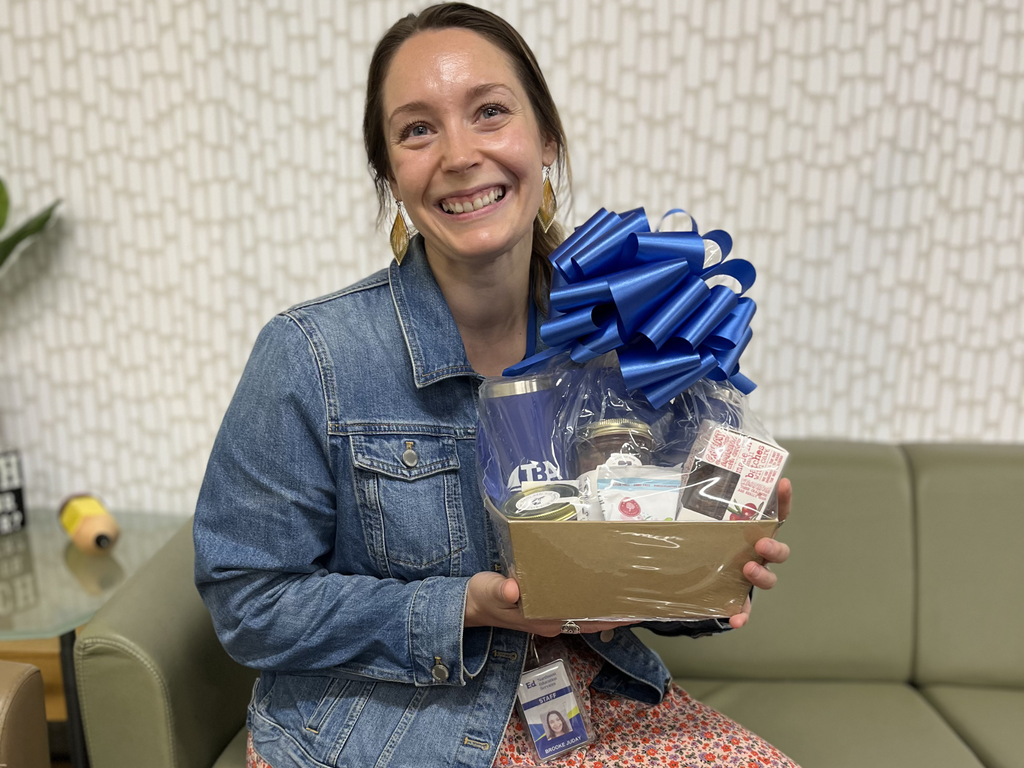 Brooke Juday sitting on a green couch, smiling and posing while holding a gift basket with a large blue ribbon. 