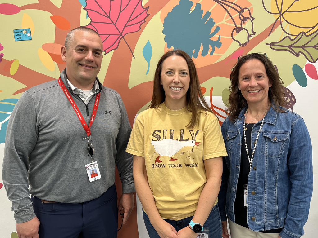 Three individuals from Suttons Bay Schools smiling and posing for a photo in front of a colorful mural. 
