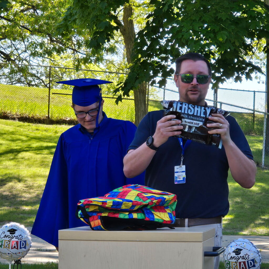 2026 Bridgeway Program graduate standing at the podium during the ceremony with a staff member who is holding a large pack of Hersey’s chocolate. 