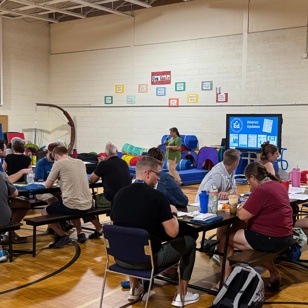 North Ed staff members sitting at lunch tables in the Bridgeway gymnasium, watching a presentation for Opening Day. 