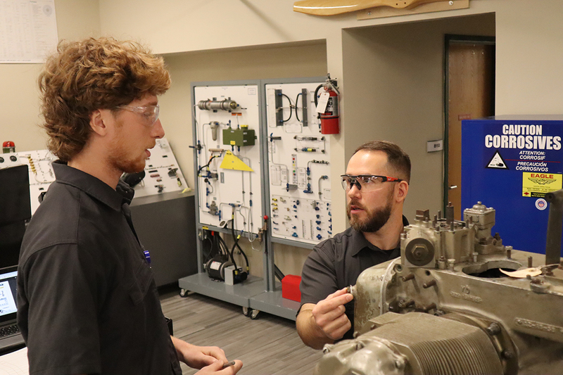 A student talks with his teacher in a school workshop. Both are wearing black shirts and wearing safety glasses. Tools are displayed in the background. 