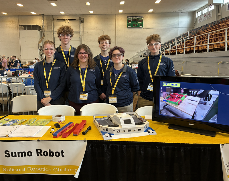 Students stand by a table with a robot and TV screen. They are on the floor of a large gymnasium-type building. Bleacher seating  is in the background.
