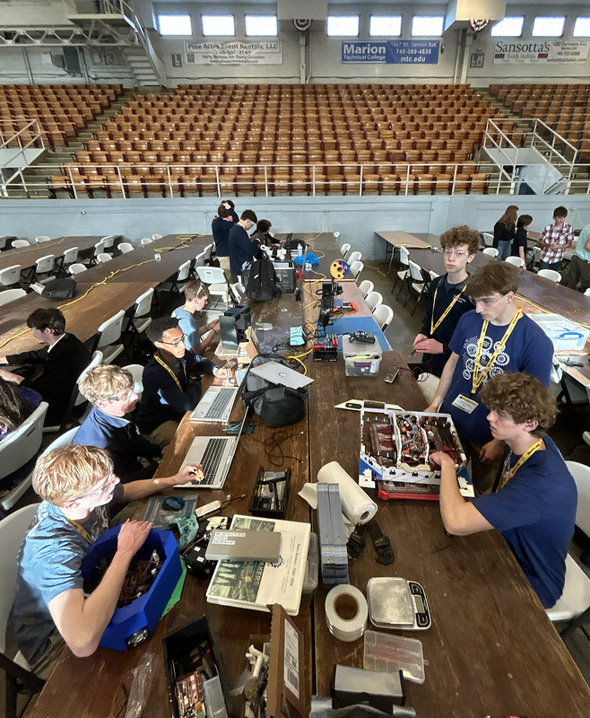 Students sit at a long table with robots and equipment. They are on the floor of a large gymnasium-type building. Bleacher seating  is in the background.