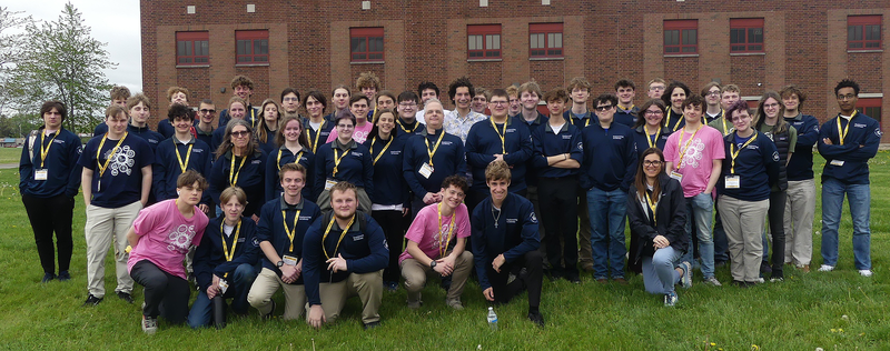 A large group of students on a school trip pose on the lawn in  front of a brick building. 