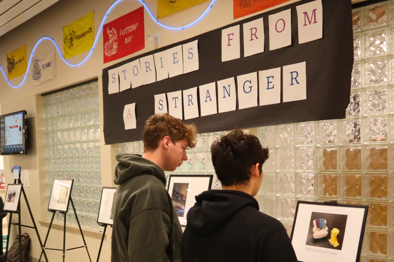 Two students view a photo placed on an easel during the Stories from a Stranger gallery event.  A banner with the name of the event is in the background .