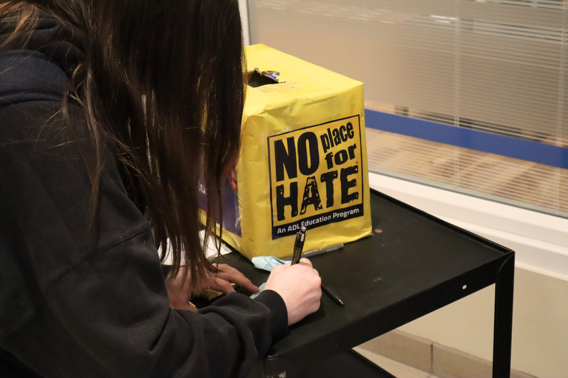 A student writes something on a piece of paper next to a small yellow box with the words No Place For Hate on it. 