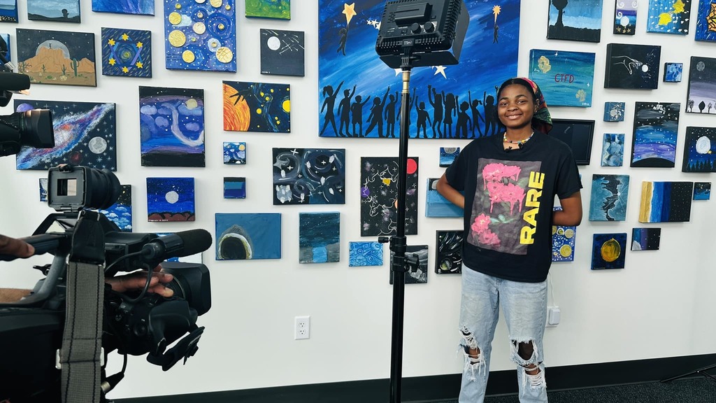 Photo of a student standing in front of the artwall, being interviewed by the news.