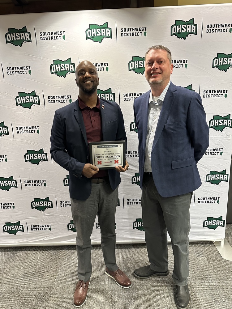 A medium-shot photo shows two men standing in front of a white backdrop featuring the "OHSAA" and "Southwest District" logos. On the left, a man with a beard, wearing a maroon polo under a navy blazer and grey trousers, proudly holds a plaque that reads "Chuck Richardson, Norwood High School." To his right stands a taller man in a light-patterned button-down shirt, a navy blazer, and grey trousers. Both are smiling at the camera during the 2026 SWOADA awards ceremony.