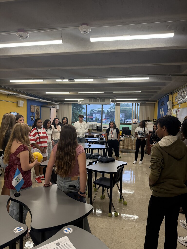 Students and teacher standing in a circle passing a ball around as they introduce themselves in Spanish.