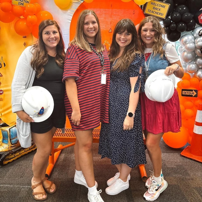 Fourth grade teachers stand smiling in front of a construction themed backdrop