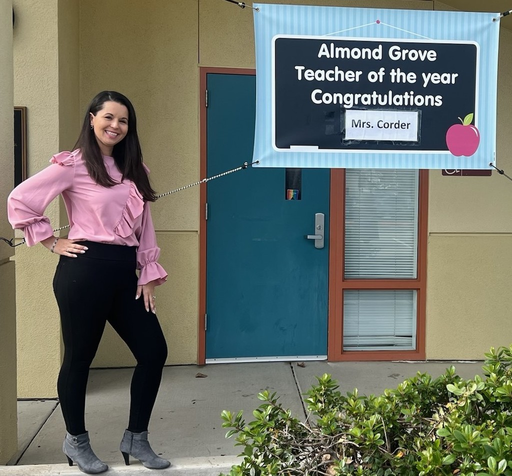Teacher standing next to a sign that says almond grove teacher of the year congratulations mrs. corder