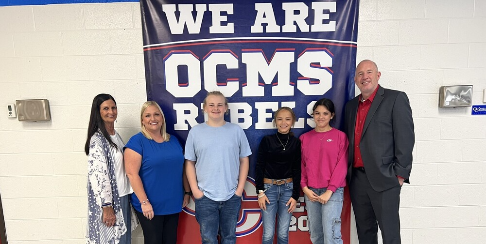 Six people pose together in front of an “OCMS Rebels” banner.